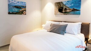 Une chambre à coucher avec un grand lit, deux tableaux représentant des paysages côtiers et une table de chevet avec une lampe.