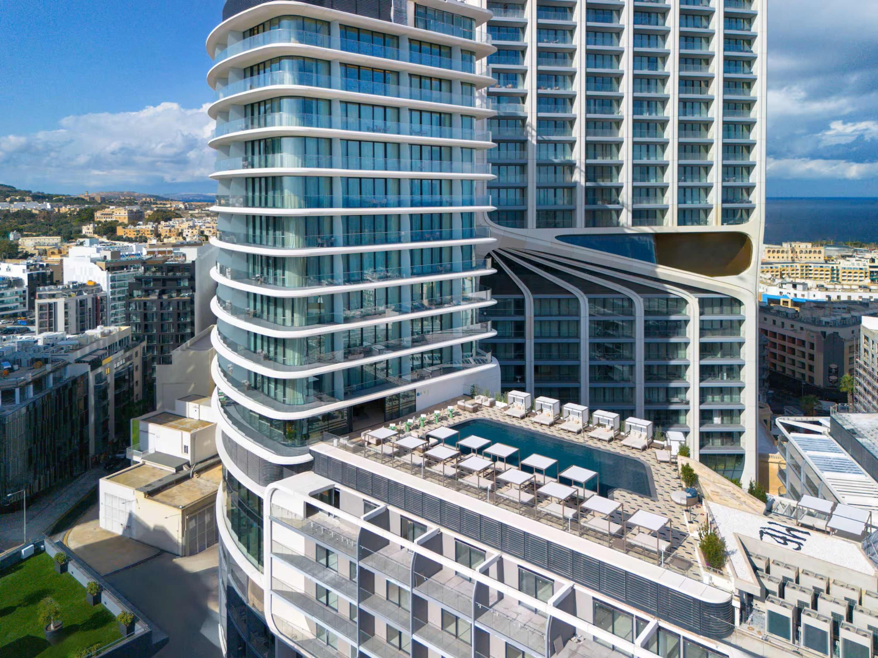A modern high-rise hotel with a curved facade, a rooftop pool area with sun loungers, and a view of the city and sea.