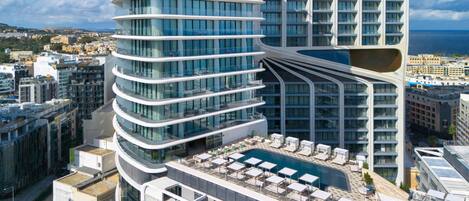 A modern high-rise hotel with a curved facade, a rooftop pool area with sun loungers, and a view of the city and sea.