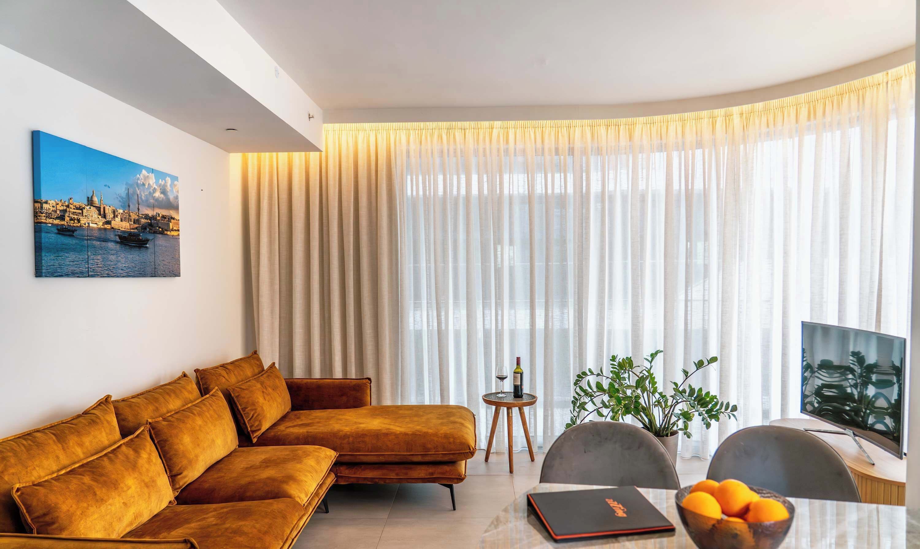 A modern living room with a sofa, a dining table with chairs, a TV, and a window with sheer curtains.