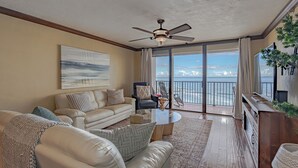 A spacious living room with a large window, a ceiling fan, a sofa, a coffee table, and a view of the ocean.