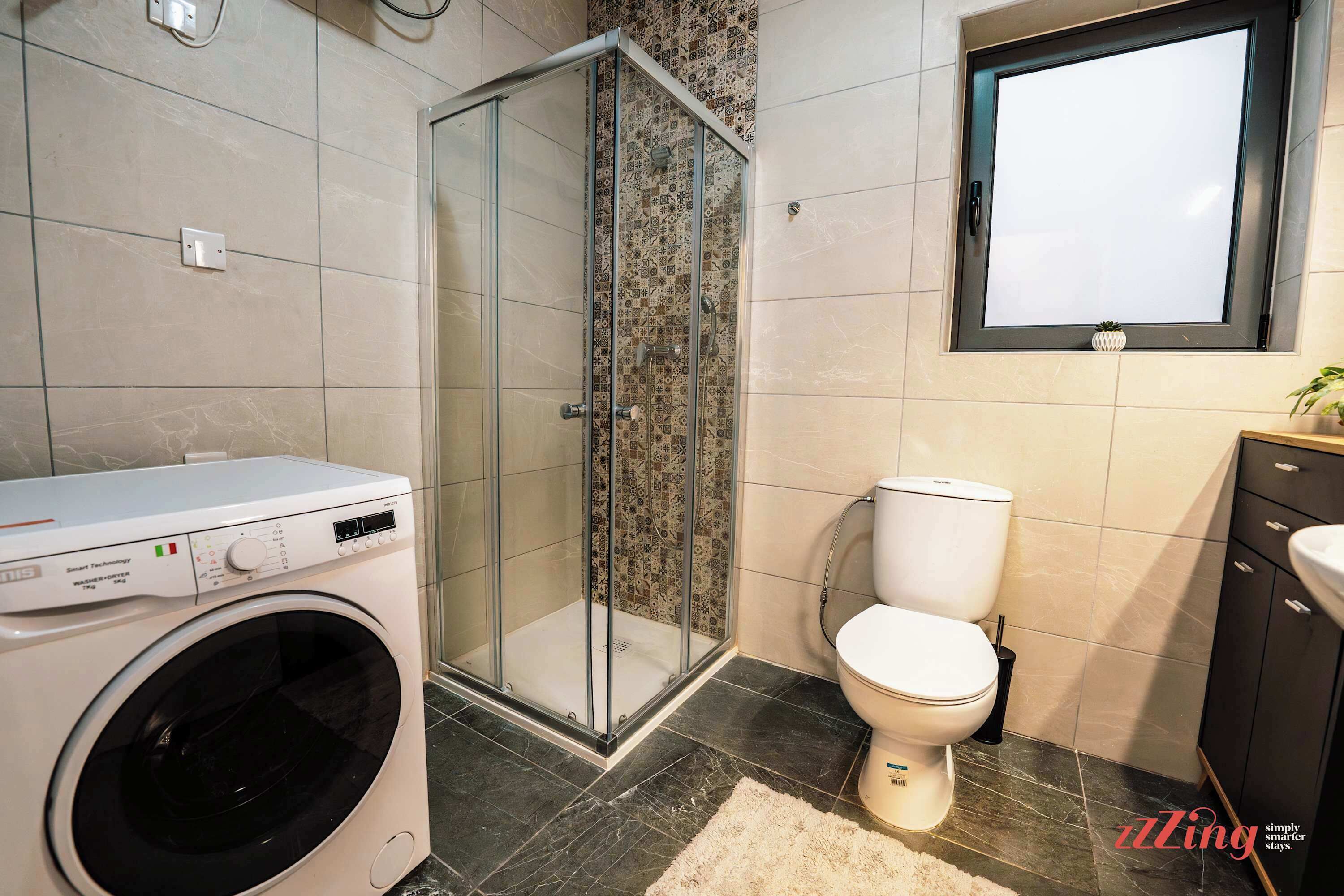 Un baño moderno con lavadora, cabina de ducha con vidrio, inodoro y lavamanos.