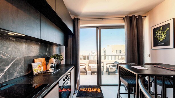 A modern kitchen with dark cabinetry, a black countertop, and a dining area with a table and chairs. - Msida's Most Loved Stylish Duplex Retreat (Tal-Pietà)