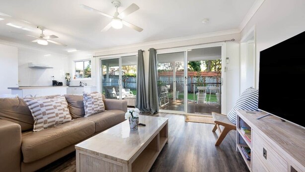 A modern living room with a sofa, coffee table, and TV stand. There is a sliding glass door leading to a patio with outdoor furniture.