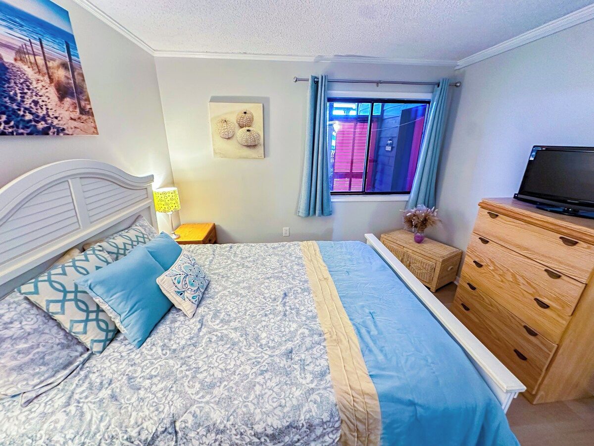 A bedroom with a bed, a dresser, a nightstand, a lamp, a window with curtains, and a painting on the wall.