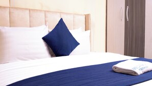 A neatly made bed with a blue bedspread and pillows, a folded towel with text, and a wooden wardrobe in the background.