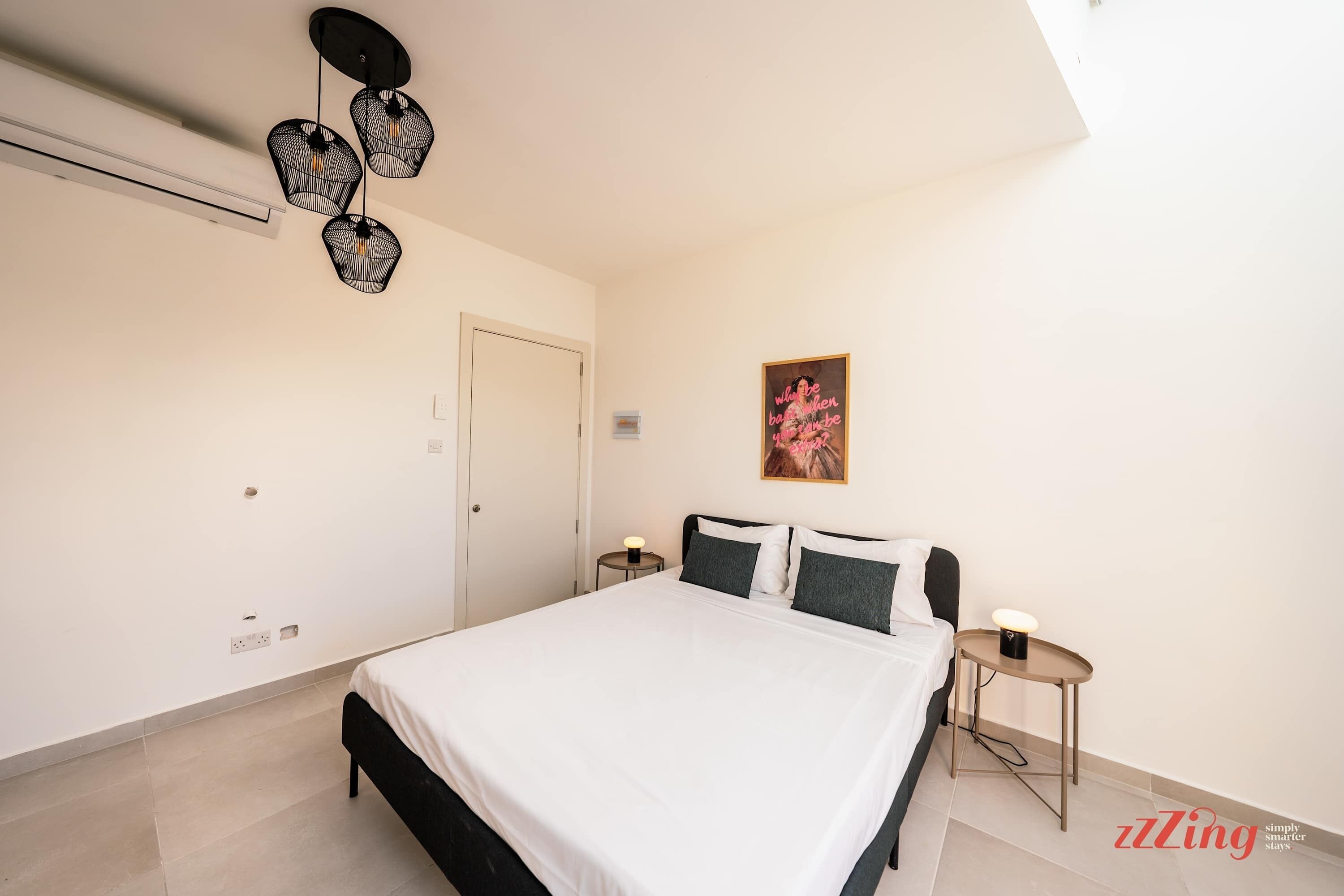 Une chambre à coucher comprenant un lit, des tables de chevet, un luminaire au plafond, une porte et une œuvre d’art accrochée au mur.