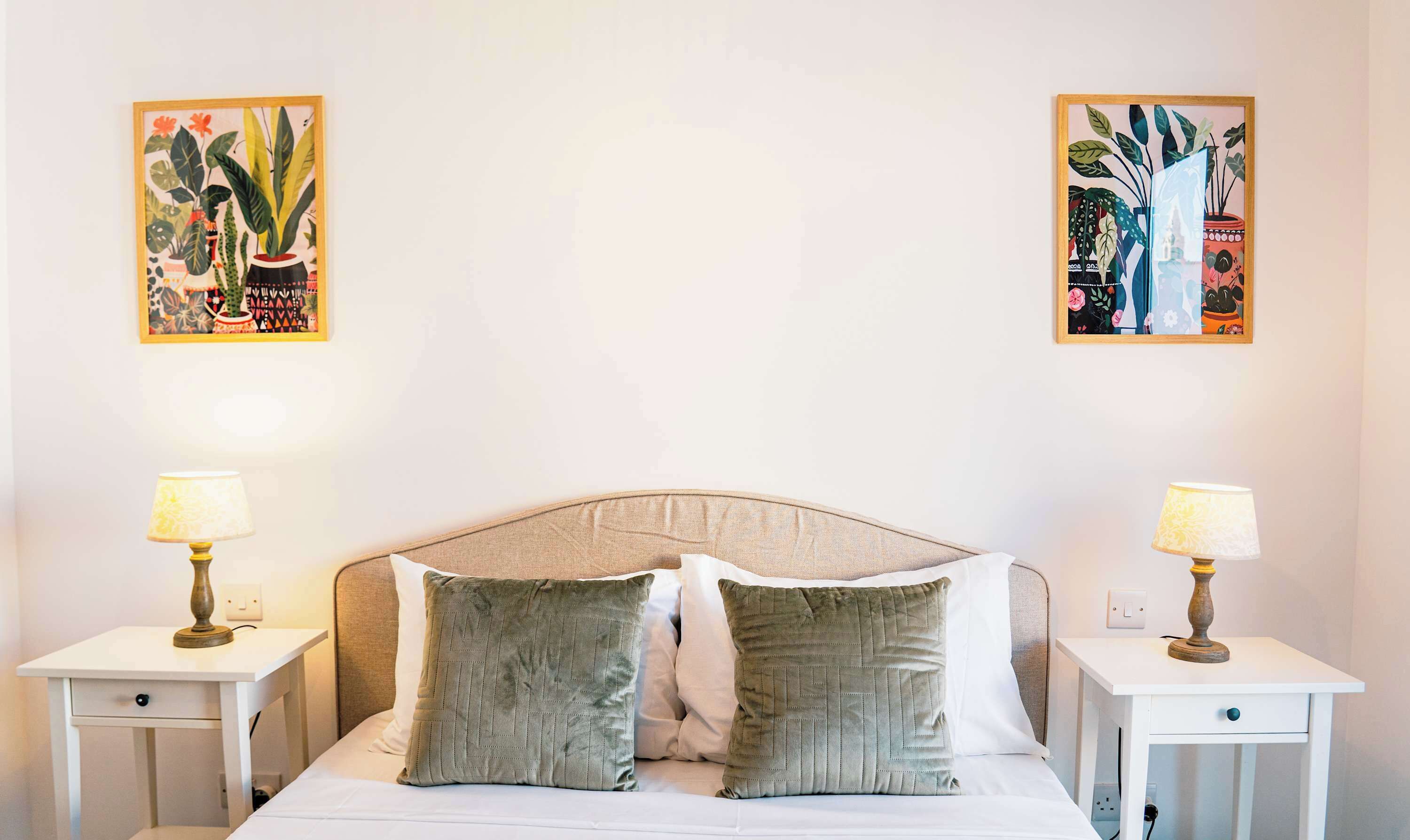 A bedroom with a bed, two nightstands, two lamps, and two wall-mounted paintings.