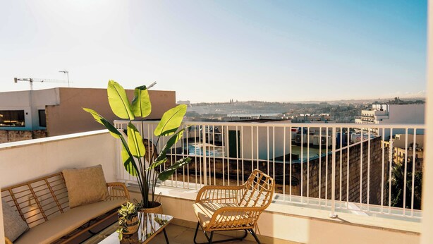 Ein Balkon mit einer Couch, einem Korbstuhl und einem Topfpflanzen, mit Blick auf eine Stadtlandschaft mit Gebäuden und einem Gewässer.