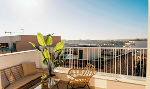 Un balcon agrémenté d’un canapé, d’un fauteuil en osier et d’une plante en pot, offrant une vue sur un paysage urbain avec des bâtiments et une étendue d’eau.