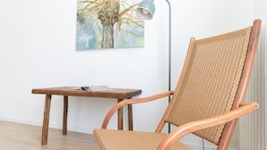 A wicker chair with a wooden frame, a small wooden table with a magazine, and a floor lamp against a white wall.
