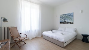 A bedroom with a bed, a chair, a table, a radiator, and a painting on the wall.