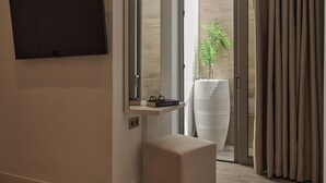 Ein modernes Badezimmer mit einem großen Spiegel, einem an der Wand befestigten Flachbildfernseher, einem kleinen Regal mit Büchern und Brillen sowie einer üppigen Topfpflanze.