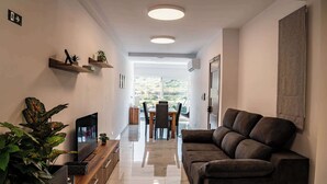 A modern living room with a flat-screen TV, a sofa, a dining area with chairs, and a view of the outdoors.