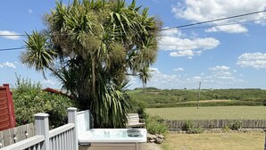 Une terrasse en bois avec un jacuzzi, un canapé en osier et une vue sur un champ verdoyant et des arbres.