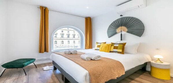 A hotel room with a bed, a small stool, a bedside table with a lamp, a window with curtains, and a view of a building outside.