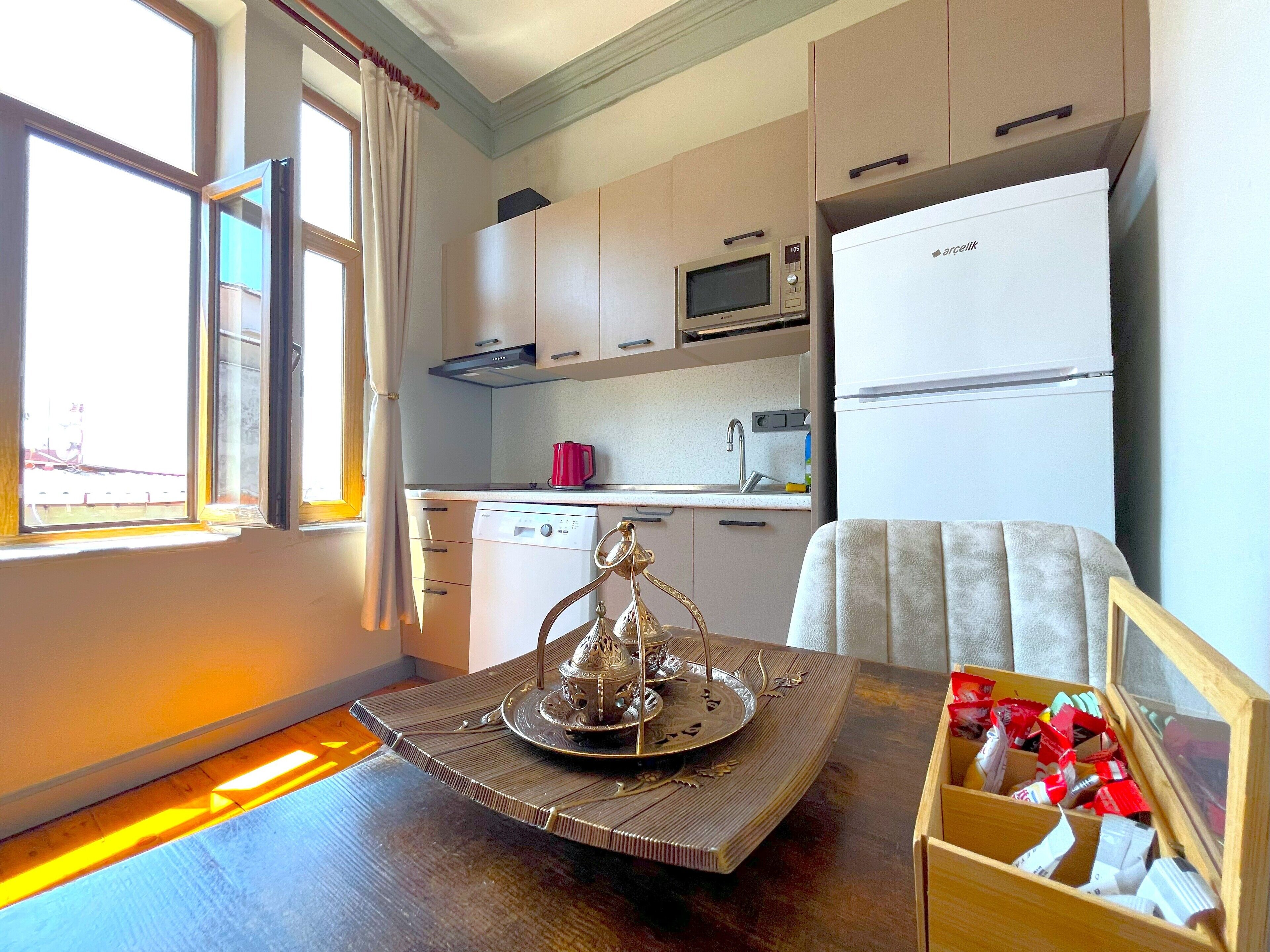 A compact kitchen with a microwave, a refrigerator, and a dishwasher. There's a window with curtains, a wooden table with a decorative tray, and a chair with a cushion.