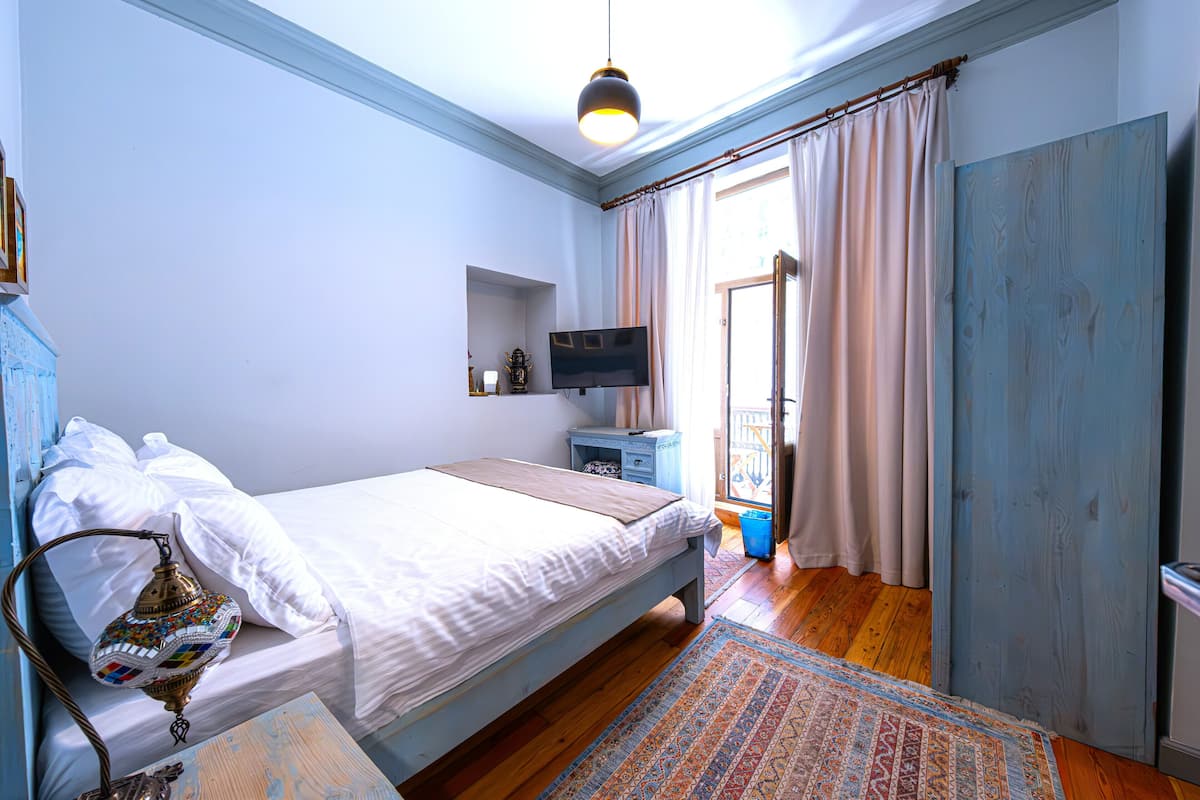 A bedroom with a bed, a television, a nightstand, a lamp, and a view of the outdoors.