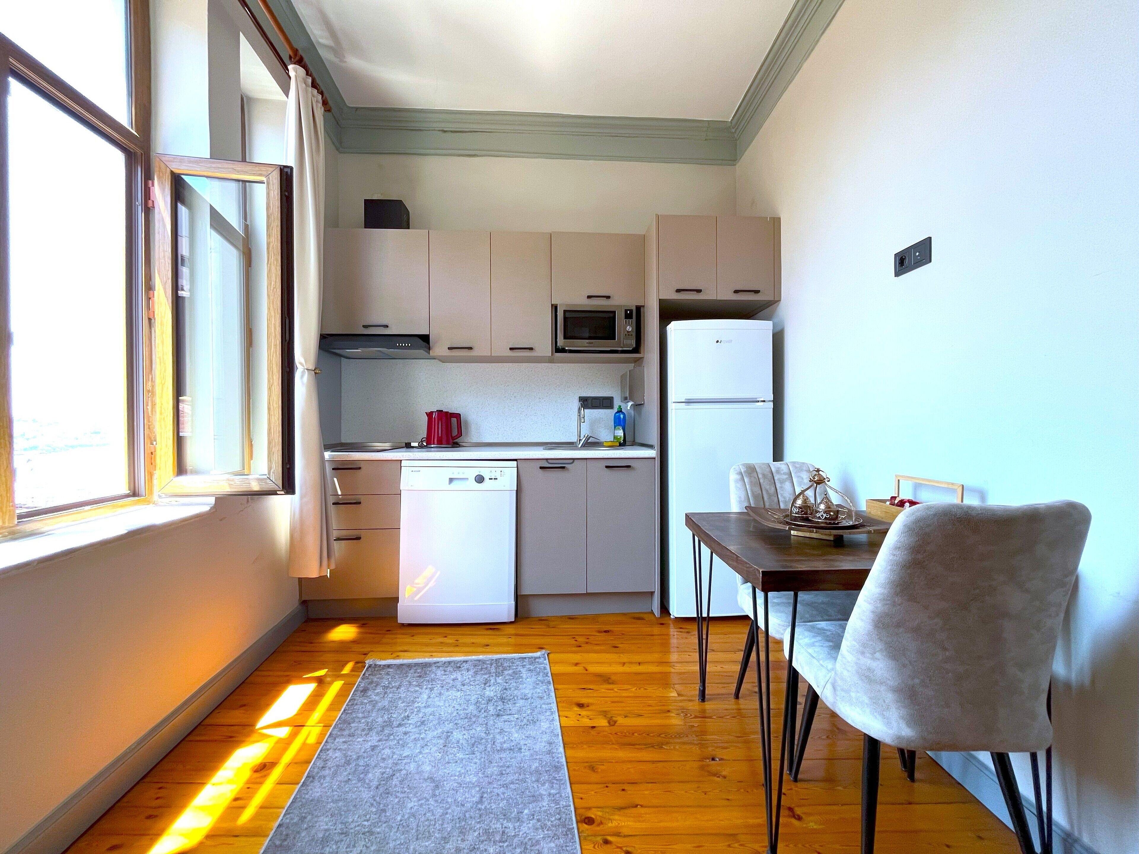 A compact kitchen with a dishwasher, microwave, and refrigerator. There is a dining table with chairs and a window with curtains.