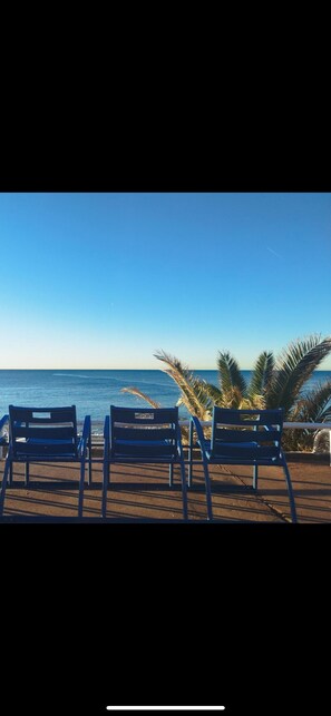 A beach view with three blue chairs facing the ocean, palm trees, and a clear sky. - The Sun of Nice (Nice)