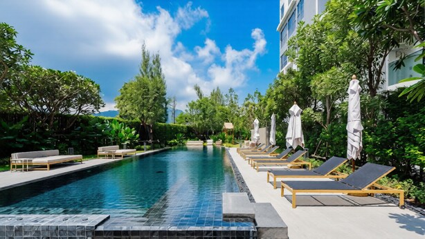 A swimming pool with lounge chairs and umbrellas, surrounded by greenery and a building.