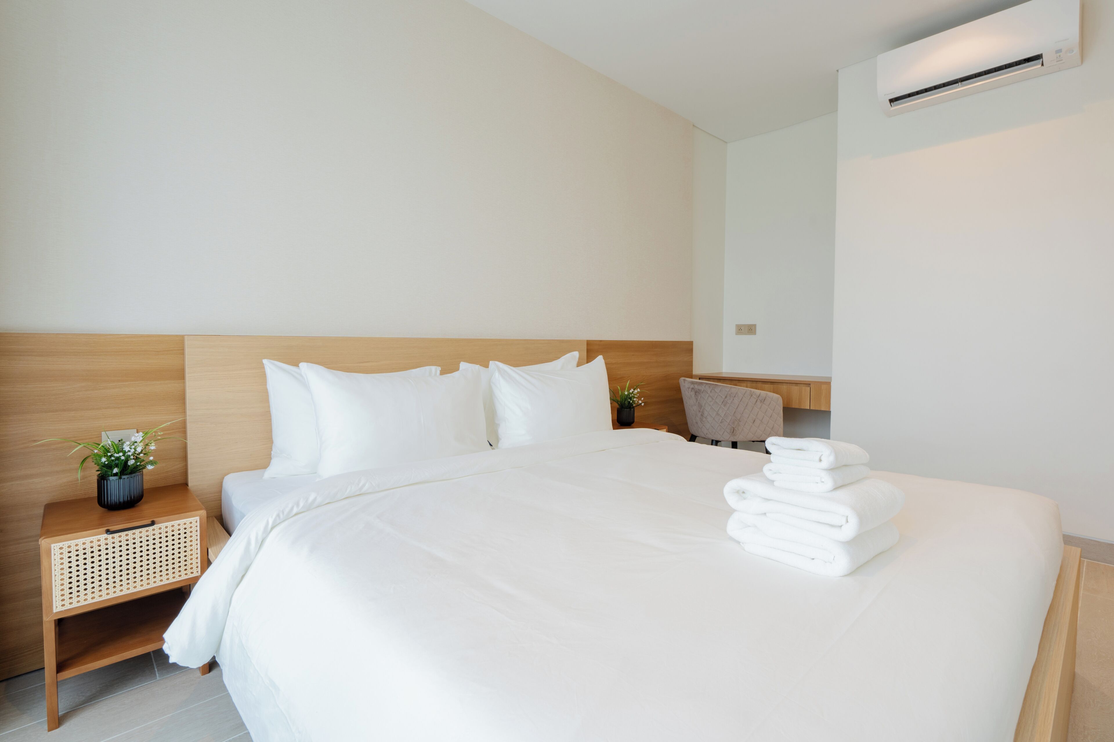 A hotel room with a large bed, a wooden headboard, a nightstand with a plant, and a wall-mounted air conditioner.