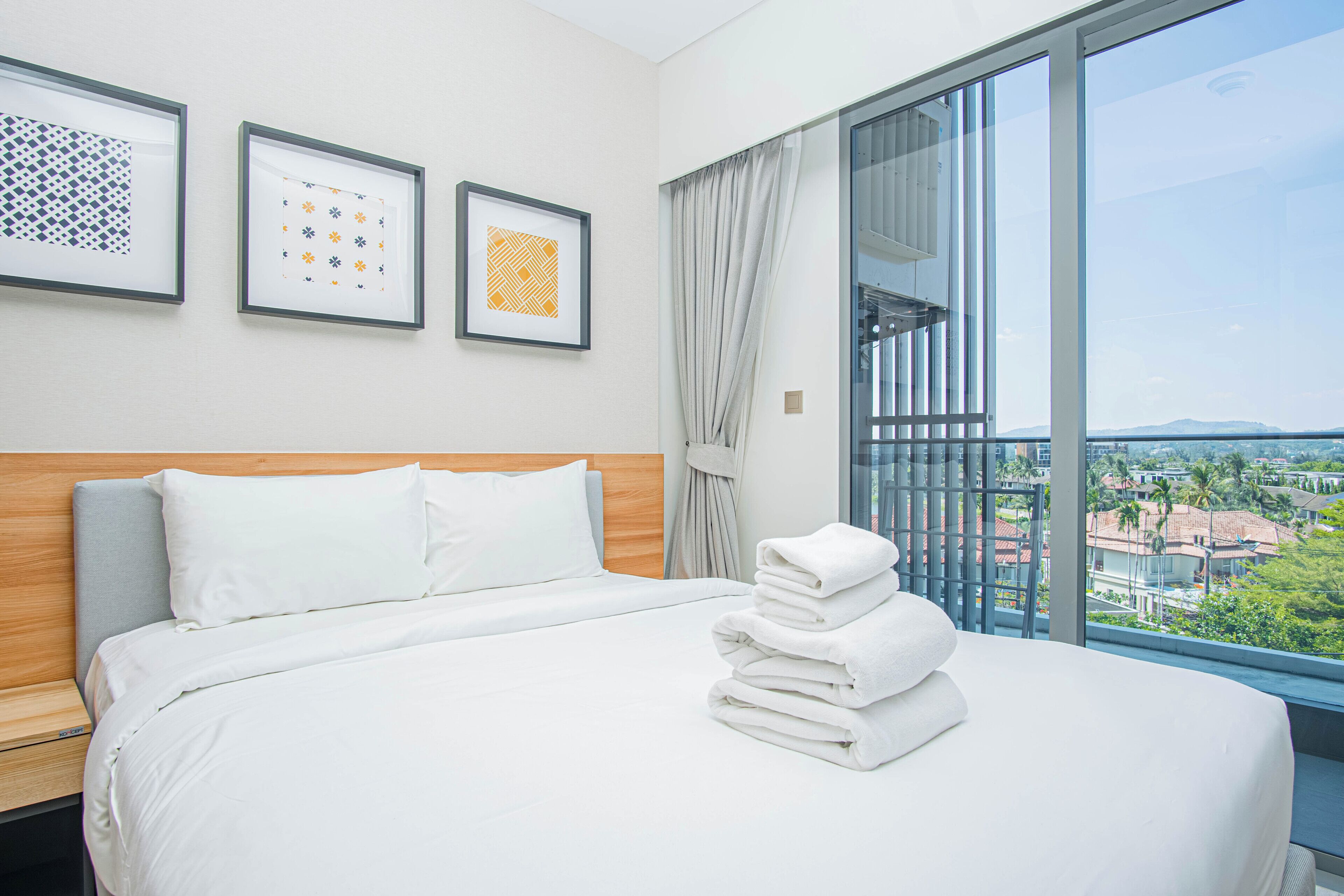 A hotel room with a bed, a view of the city, and framed artwork on the wall.