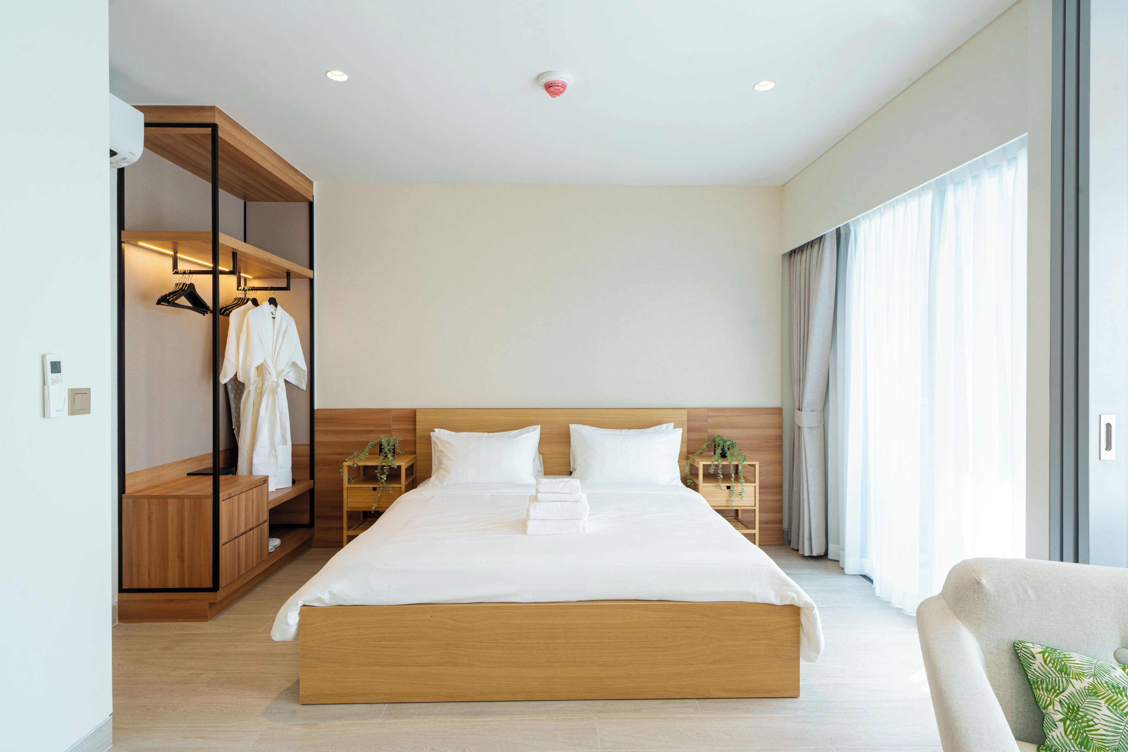 A modern hotel room with a large bed, wooden headboard, built-in wardrobe, bedside tables, and a large window with curtains.