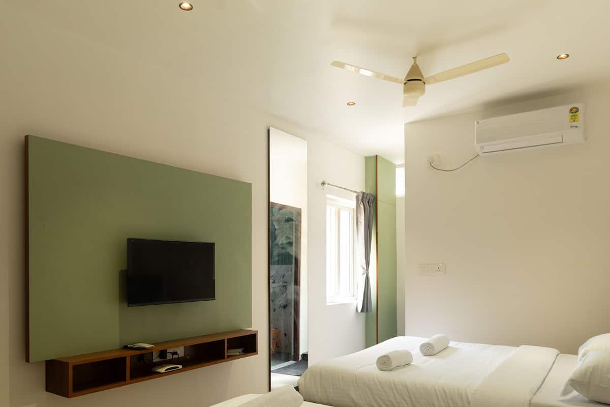 Habitación de hotel con dos camas, un televisor de pantalla plana montado en una pared verde, un ventilador de techo y un equipo de aire acondicionado.
