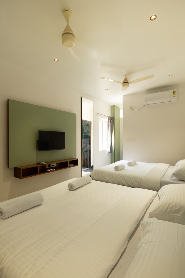 A hotel room with two beds, a flat-screen TV mounted on a green wall, a ceiling fan, and an air conditioning unit.