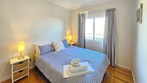A bedroom with a bed, bedside lamps, a window with curtains, and a small bedside table with a lamp.