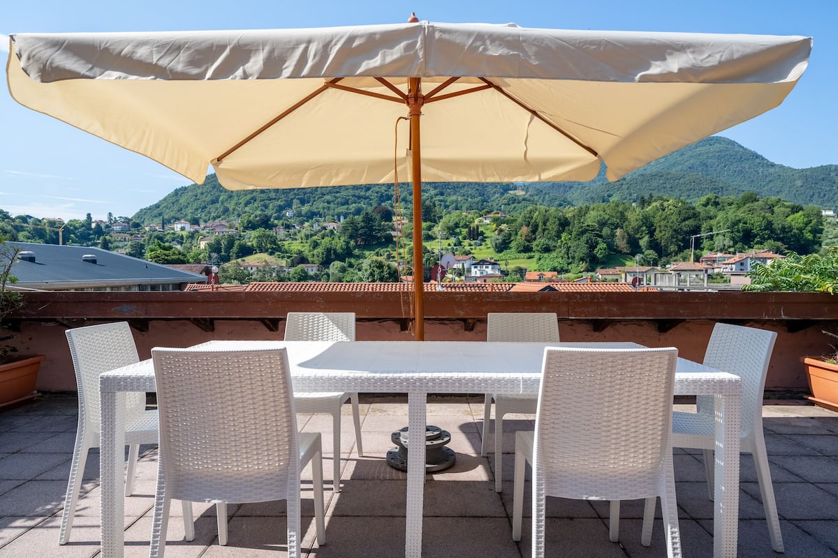 A terrace with a table and chairs under a large umbrella, overlooking a mountainous landscape with houses.