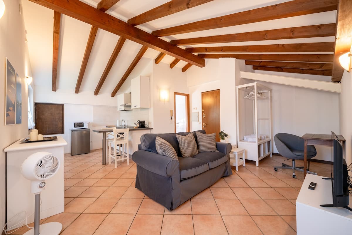 A cozy living room with a grey sofa, wooden beams, a white fan, and a small kitchen area in the background.