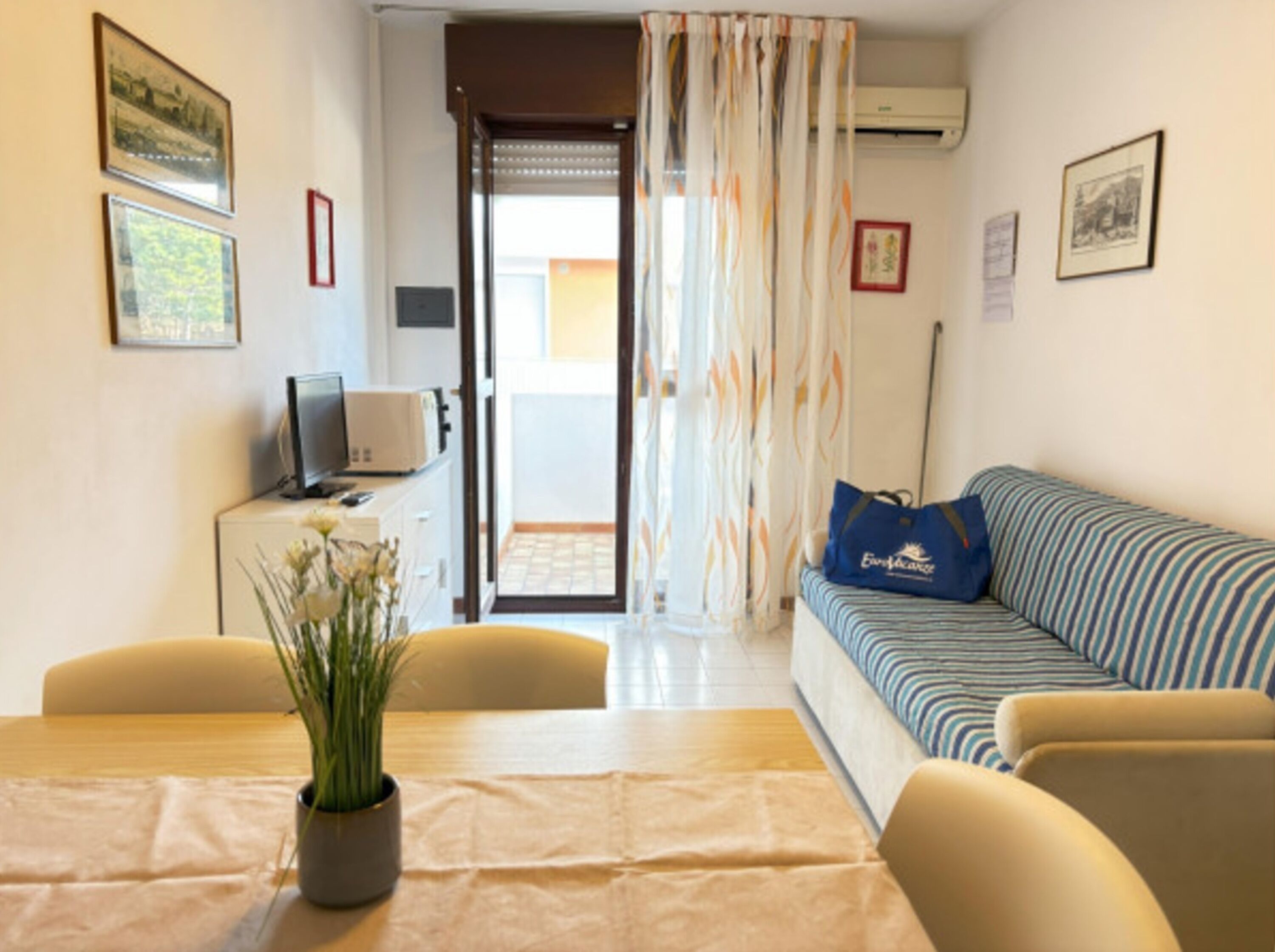 Una habitación con un sofá a rayas, una mesa de comedor con una planta, un televisor y vistas a través de una puerta.