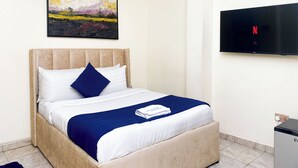 A neatly made bed with a blue and white duvet, a single blue pillow, and a white blanket. A painting of a landscape is hung above the bed. A flat-screen TV is mounted on the wall to the right.