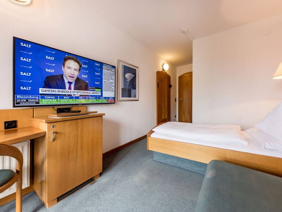 Habitación de hotel con cama, escritorio, televisión mostrando noticias financieras y un cuadro en la pared.