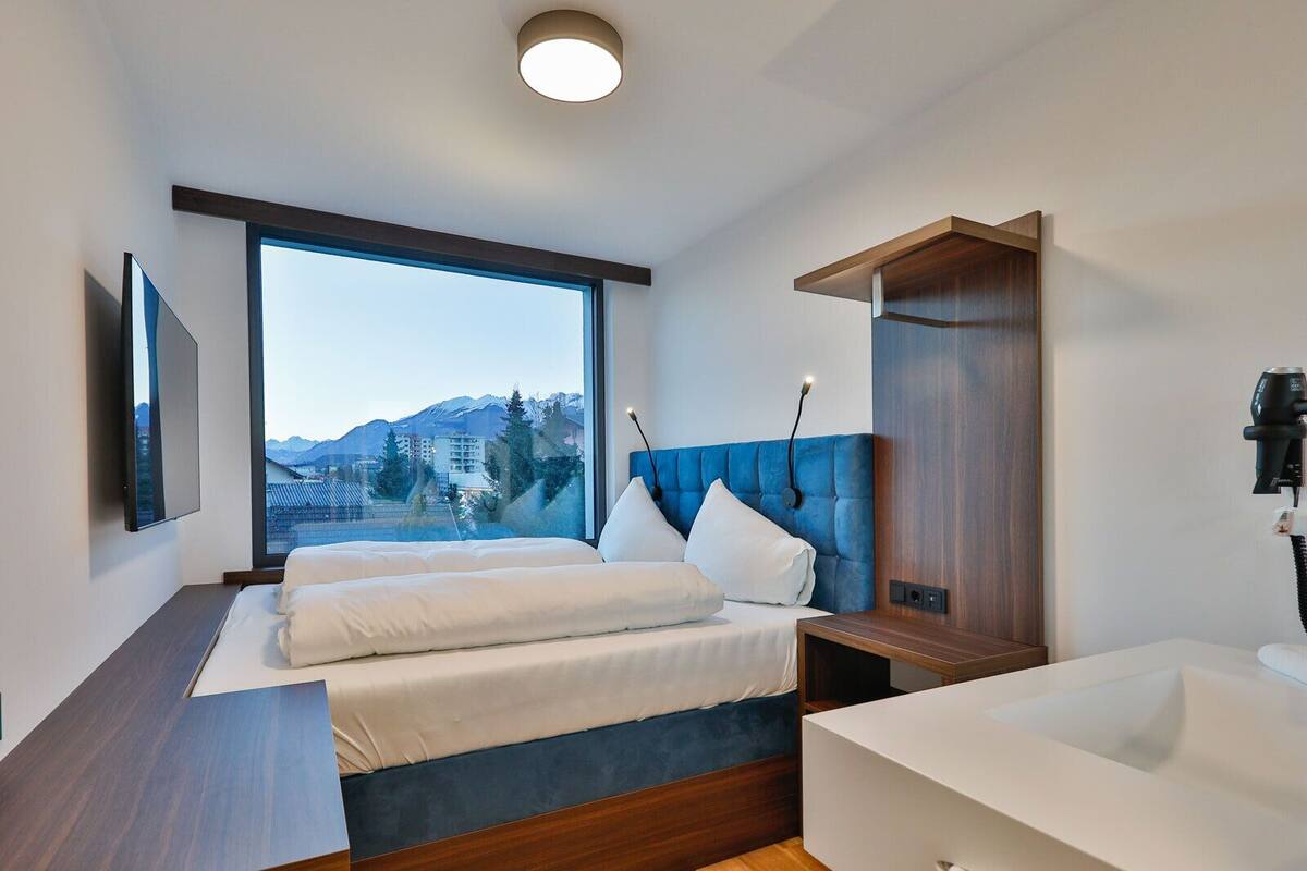 A modern hotel room with a large window, a bed with a blue headboard, a bedside table, and a bathroom with a white sink.