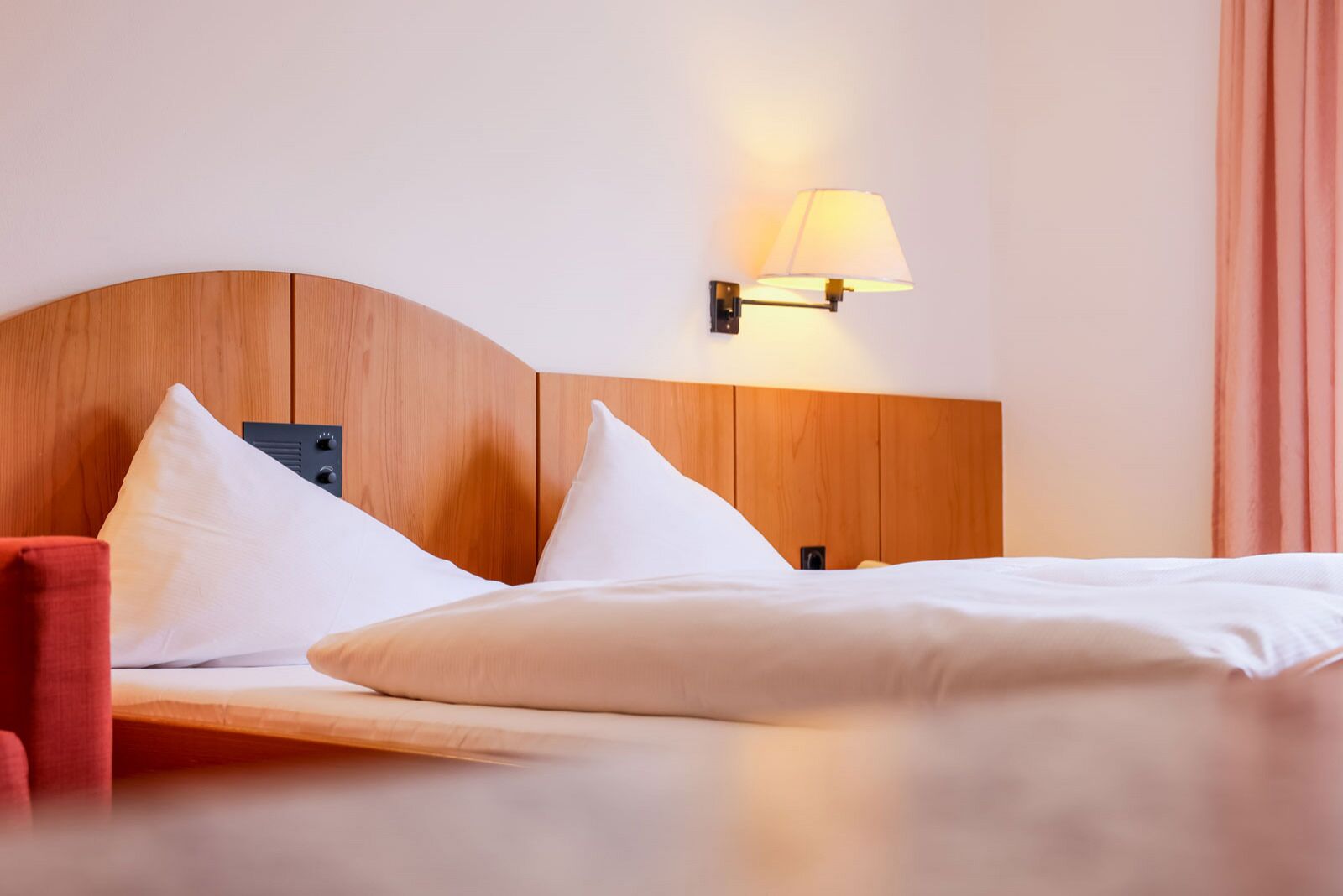 A hotel room with a wooden headboard, two white pillows, a red chair, and a wall-mounted lamp.