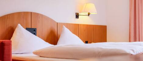 Habitación de hotel con cabecera de madera, dos almohadas blancas, una silla roja y una lámpara fijada a la pared.
