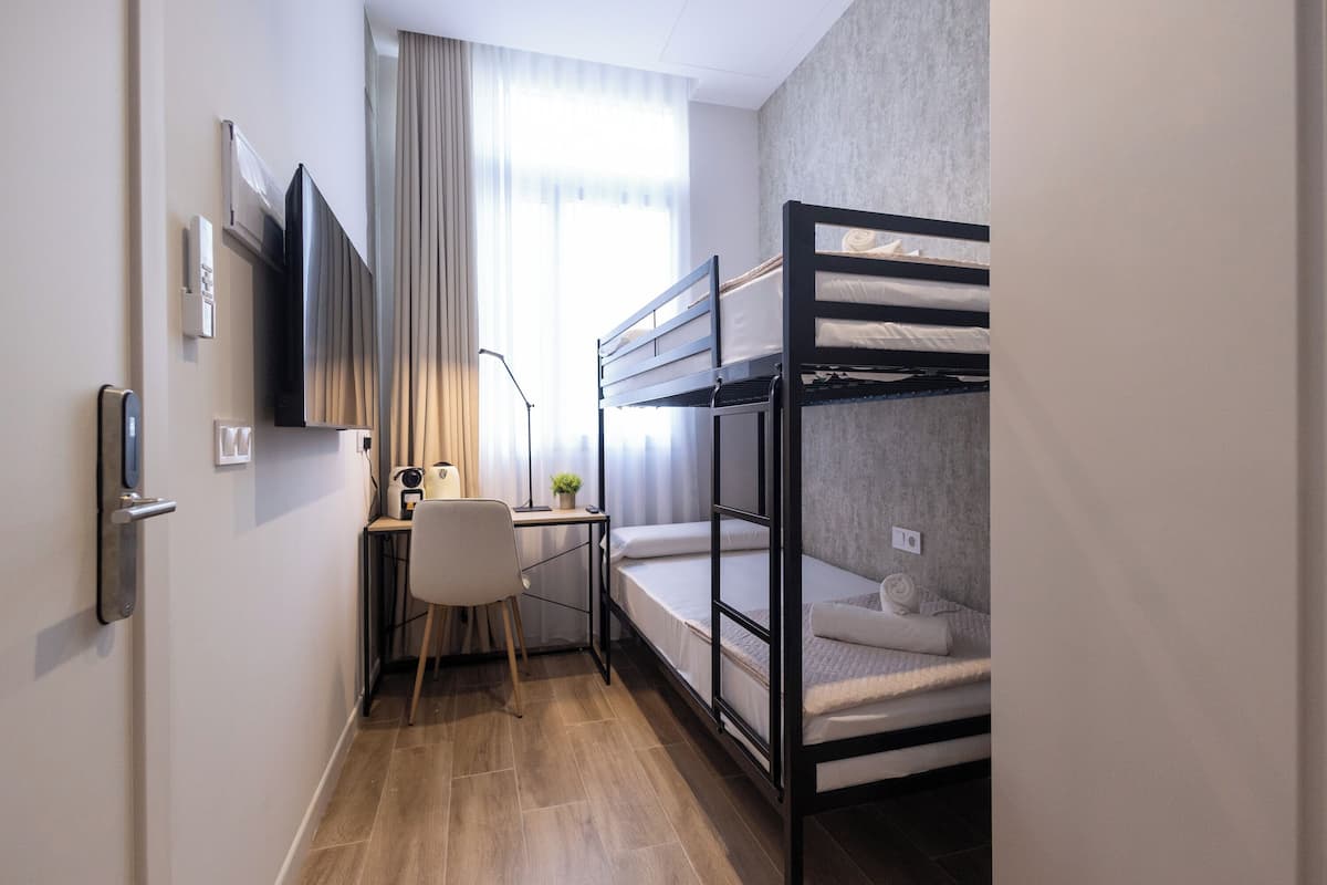 A compact hotel room with bunk beds, a desk with a chair, a small table with a plant, and a TV mounted on the wall.