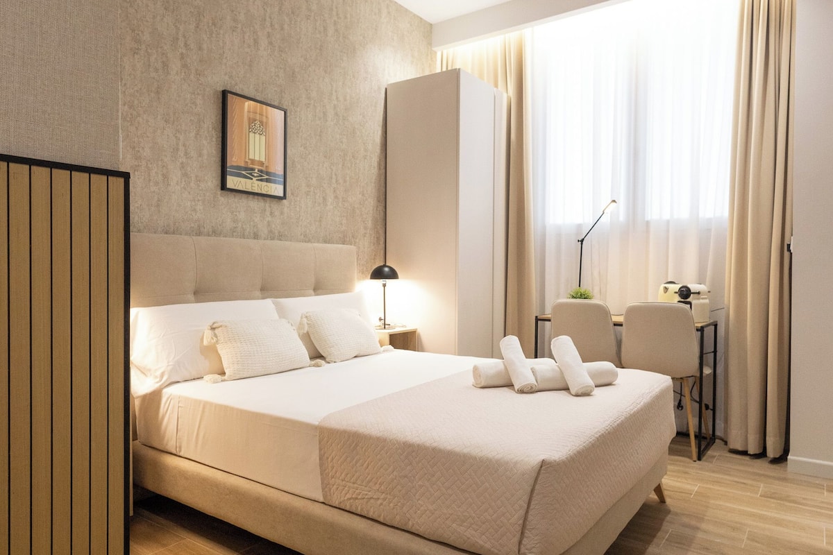 A modern hotel room with a large bed, a desk with a chair, a lamp, and a window with curtains.