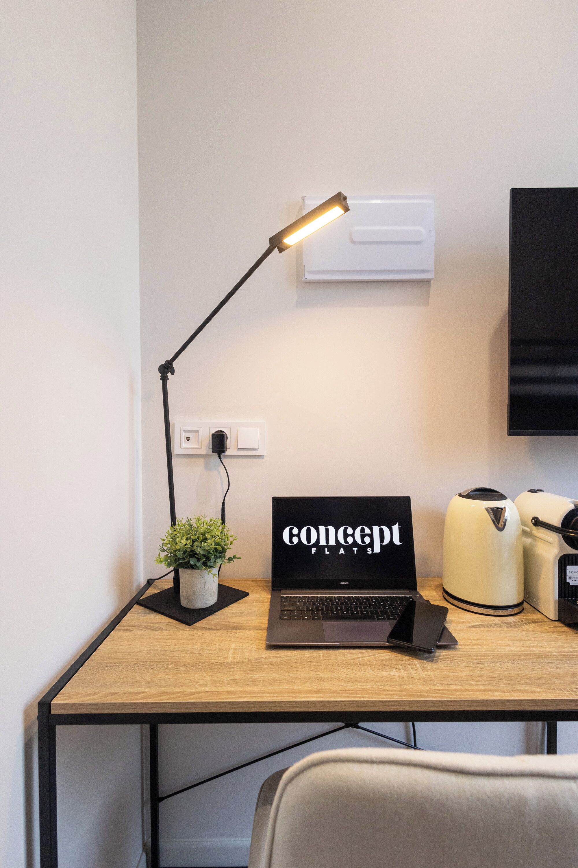 Un escritorio de madera con una computadora portátil, una lámpara, una planta pequeña en maceta, un televisor de pantalla plana y una tetera.