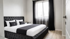 A modern bedroom with a tufted headboard, a large bed with white and black bedding, a bedside table, and a window with sheer curtains. - EF SUİT OTEL (Ankara)