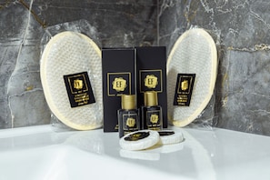 A bathroom setting with a marble wall, a white bathtub, and a set of toiletries including a bottle and a box with a gold emblem. - EF SUİT OTEL (Ankara)