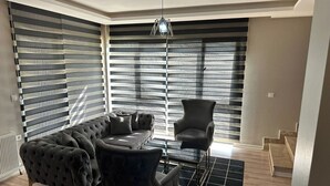 A corner room with a sectional sofa, a single armchair, a glass coffee table, and a black and white striped window blind. - EF SUİT OTEL (Ankara)