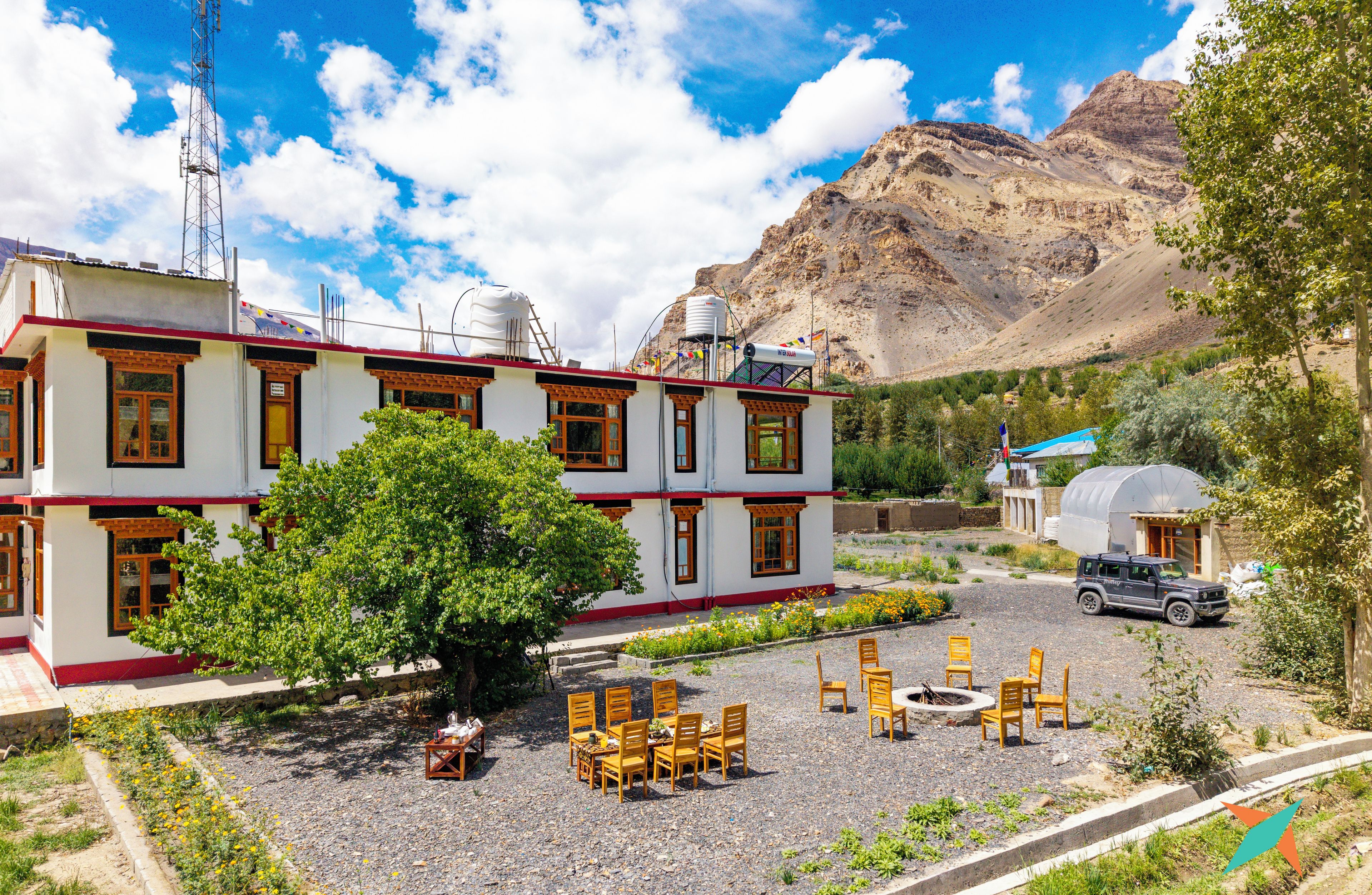 Un edificio de dos pisos con fachada roja y blanca, un patio con mesas al aire libre y un telón de fondo montañoso.