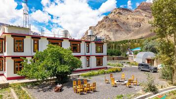 Un edificio de dos pisos con fachada roja y blanca, un patio con mesas al aire libre y un telón de fondo montañoso.