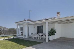 A covered patio with a dining table and chairs, a small garden area, and a white building with a tiled roof.