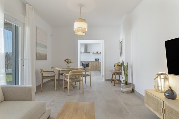 A modern living room with a sofa, dining table, chairs, a wooden coffee table, a hanging light fixture, a TV on the wall, and a kitchen in the background.