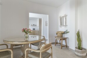 A dining area with a glass table, wicker chairs, a vase with flowers, a wooden side table, and a potted plant.